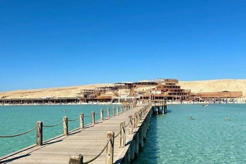 A wooden pier extending into a clear blue sea, leading to a building with multiple levels and a sandy beach in the background. 4