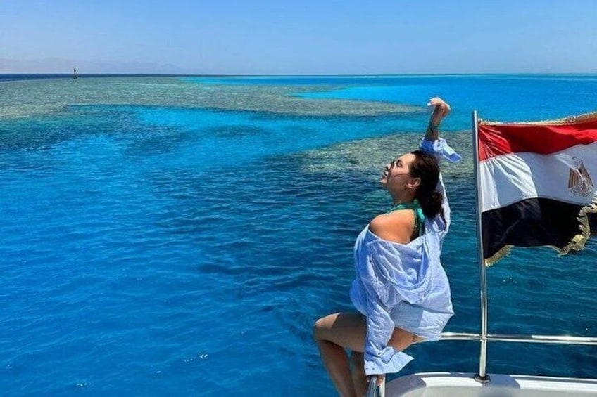 A person on a boat with a flag, clear blue sea, and distant land. 1