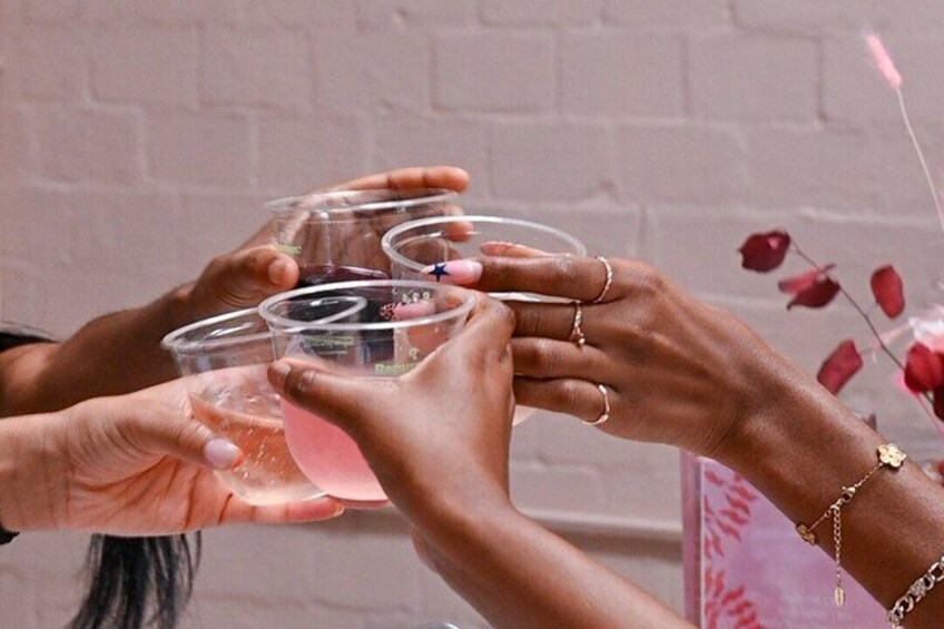Hands holding clear glasses with pink liquid, against a brick wall background. 2