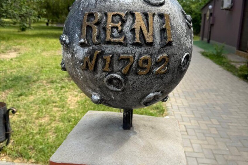 A spherical metal sculpture with 'RENI' and 'N1792' engraved on it, placed on a concrete base outdoors. 3