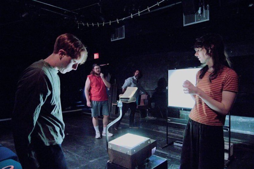 A group of people in a dimly lit room with a projector screen, a table with a laptop, and a person standing with hands clasped. 2