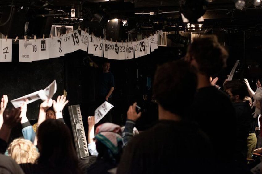 A crowd at an indoor event with a person on stage and a banner with numbers. 4