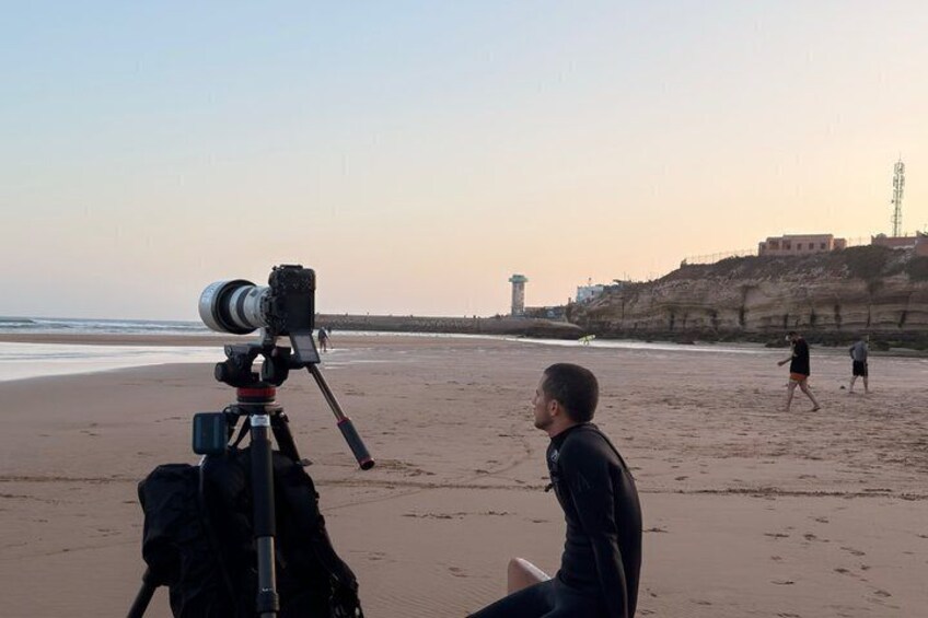 A beach with a camera on a tripod, a person sitting, and a lighthouse in the background. 1