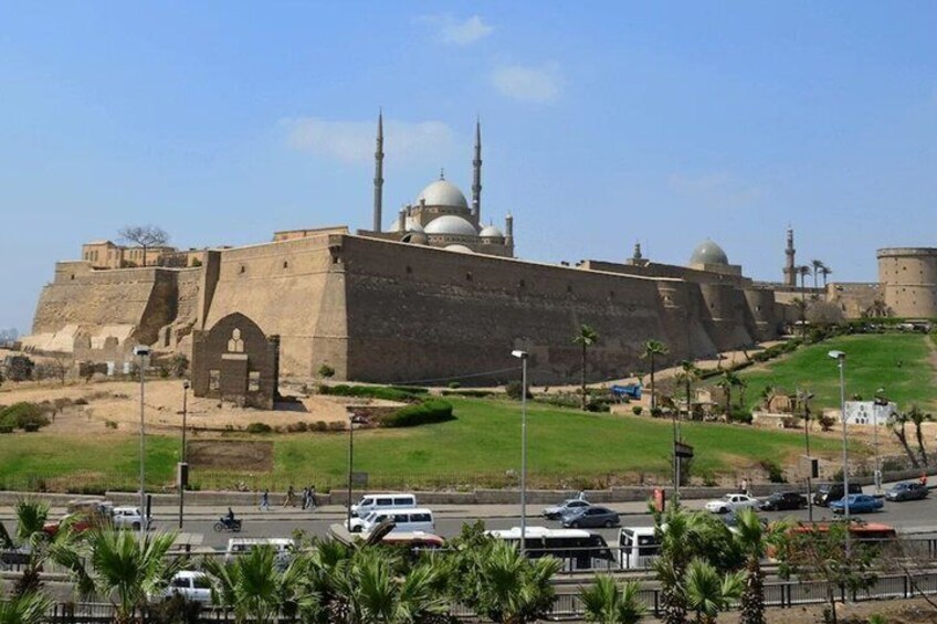 A large fortress-like structure with domed roofs and minarets, surrounded by a green landscape and a busy road. 1