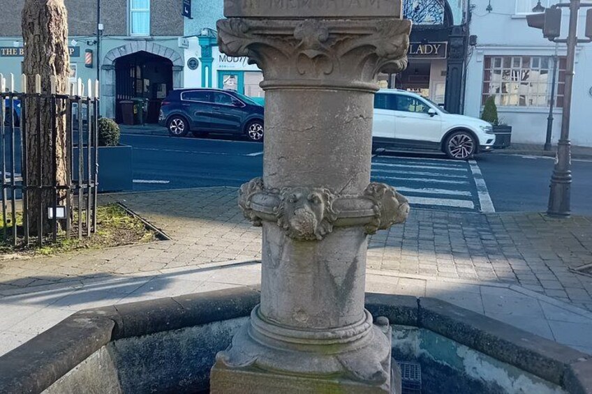 A stone fountain with a lion's head, a tree, and a street with cars and buildings in the background. 2