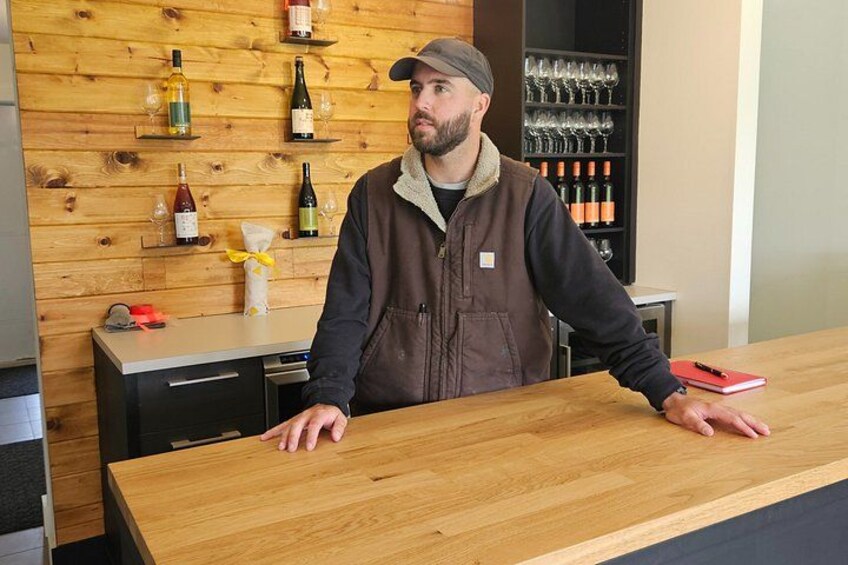 A person standing behind a wooden counter with wine bottles and glasses on shelves in the background. 3