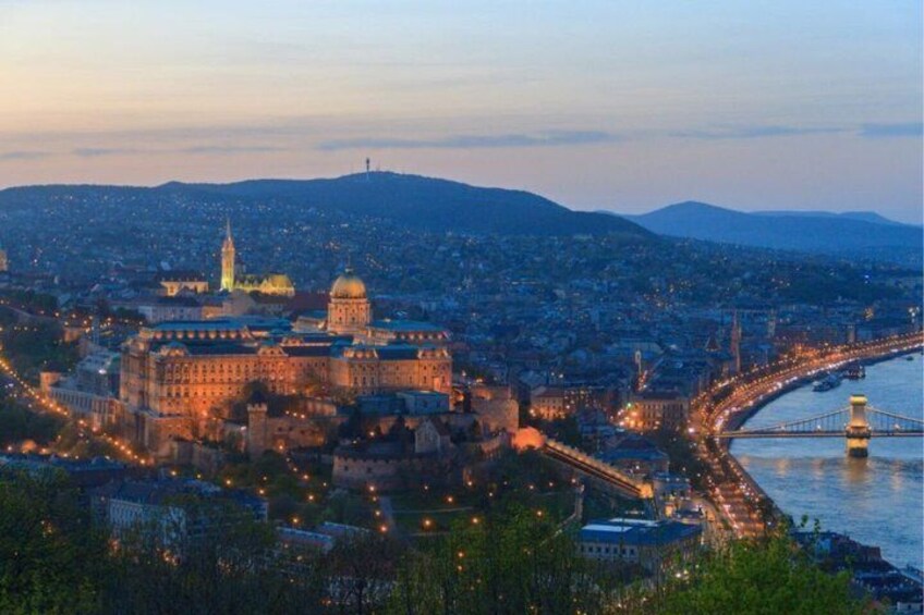 A cityscape at dusk with illuminated buildings, a prominent dome structure, and a bridge over a river. 1
