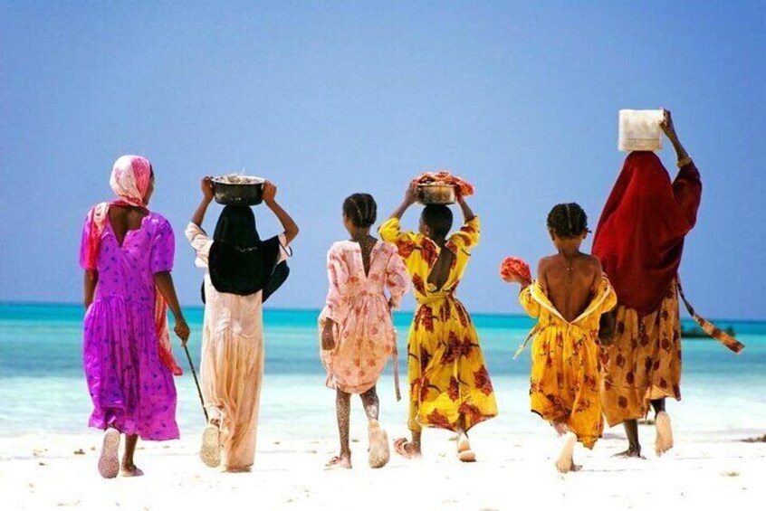 A group of people walking on a beach, some carrying items on their heads. 5