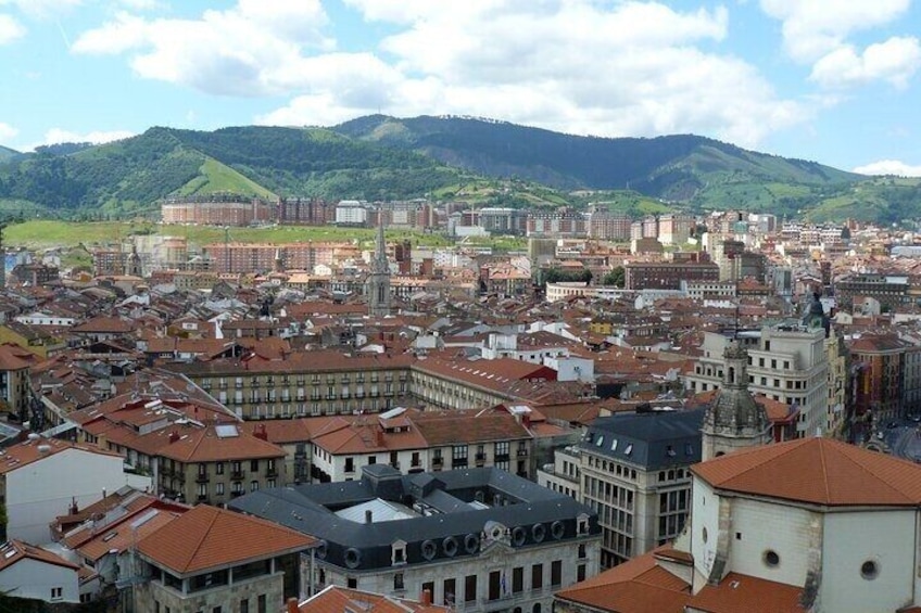 A cityscape with red-roofed buildings, a prominent church, and green hills in the background. 2