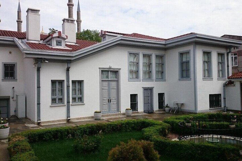A white building with red-tiled roofs, multiple chimneys, and a well-maintained garden with a circular hedge. 1