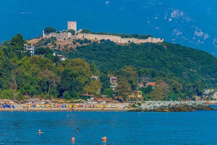 A coastal town with a historic castle on a hill, a beach with umbrellas, and calm blue waters. 5