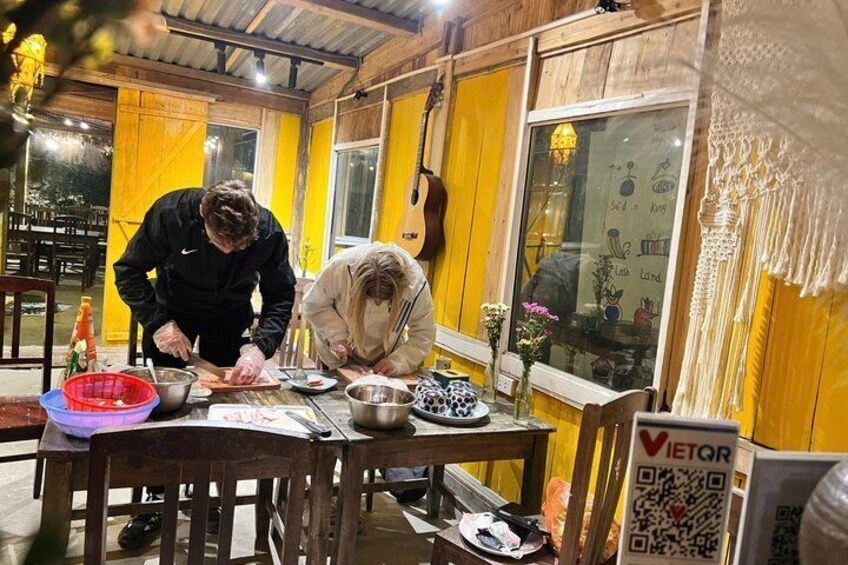 Two people preparing food at a wooden table with bowls, a cutting board, and a QR code sign. 2