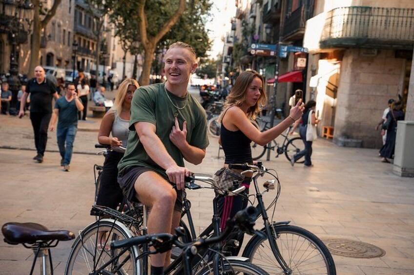 Two people on bicycles, one wearing a green shirt and the other in a black top, on a city street with pedestrians and buildings in the background. 1