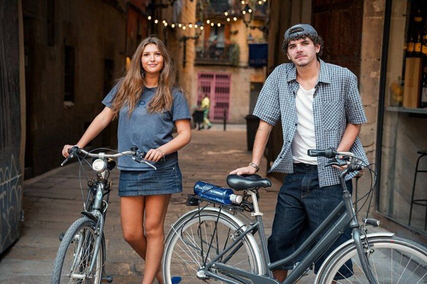 Two people standing next to bicycles in an alleyway with buildings and string lights in the background. 3