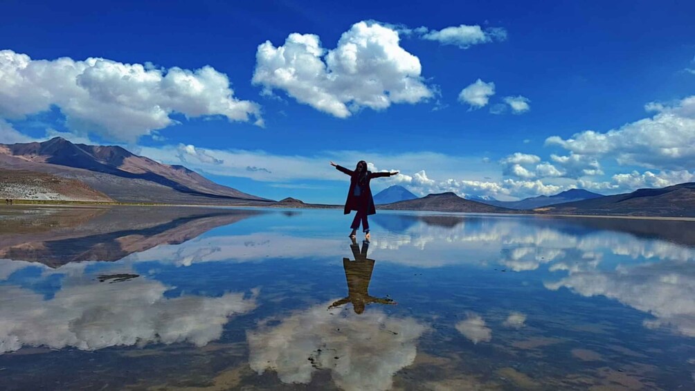 A person standing on a reflective water surface with arms outstretched, surrounded by mountains and a clear sky. 1
