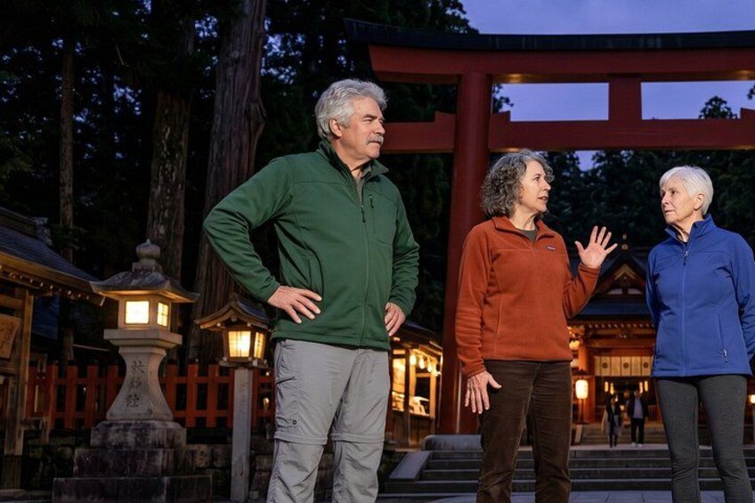 Three people standing in front of a traditional Japanese torii gate with a wooden structure and stone lanterns. 3