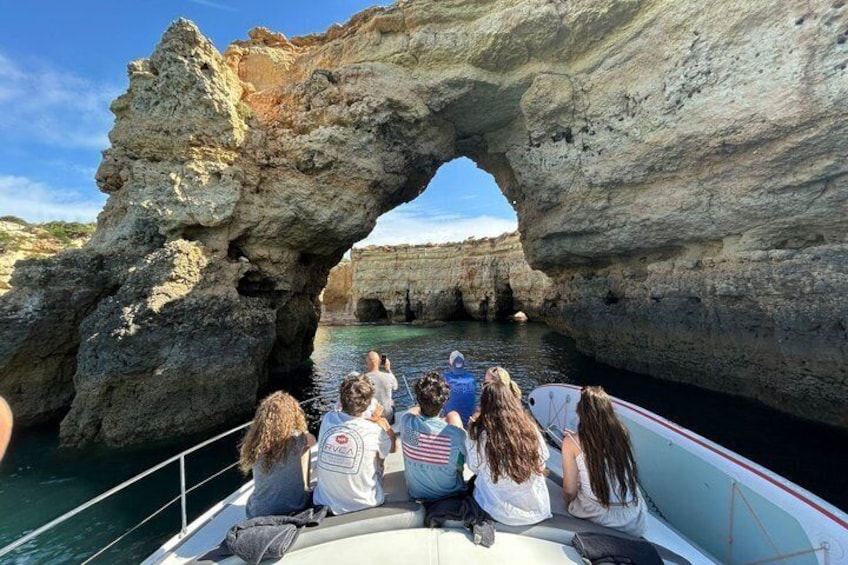 A boat with people wearing white shirts with red and blue logos, a large rock formation with an arch, and clear blue water. 1