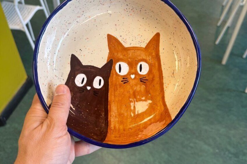 A hand holding a ceramic bowl with two painted cats, one brown and one orange, with a speckled pattern. 2