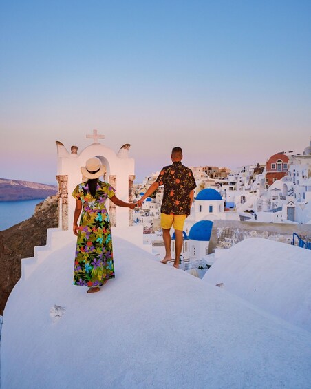 A couple holding hands on a white-washed street with blue domes, overlooking a coastal town. 1