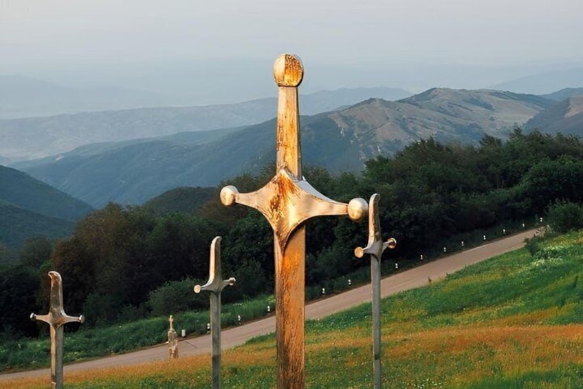 A wooden cross with a golden top, surrounded by smaller crosses, set against a hilly landscape with a road. 5