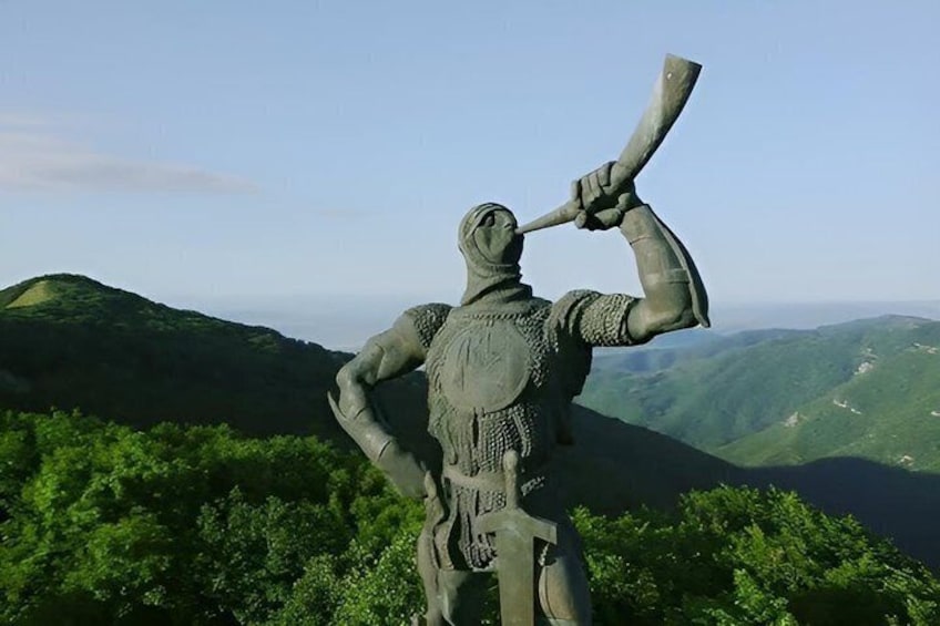 A statue of a warrior holding a spear, set against a backdrop of green hills and a clear sky. 4