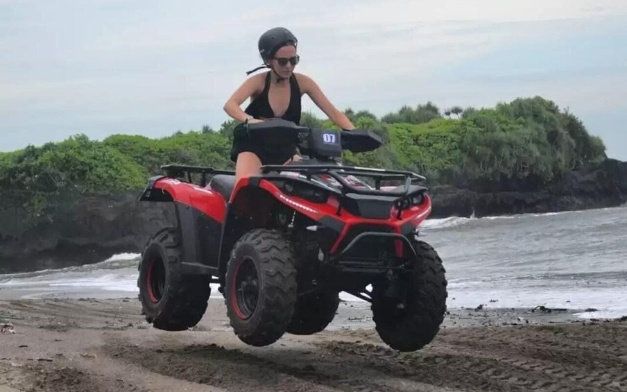 A person riding a red ATV on a beach with ocean and greenery in the background. 1