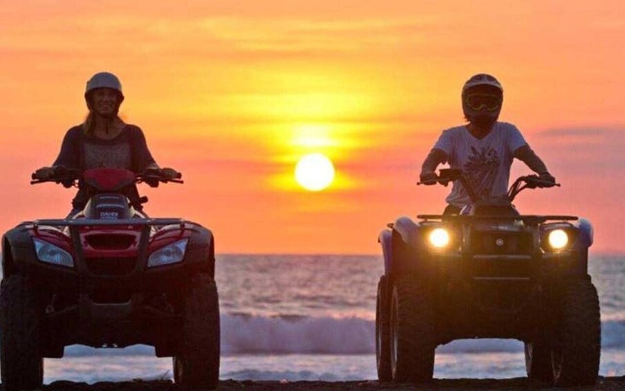 Two people riding ATVs on a beach at sunset. 1