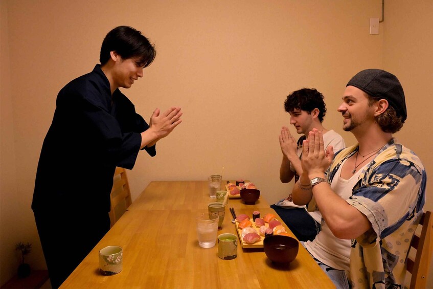 A wooden table with sushi, glasses, and a person standing with hands together. 2