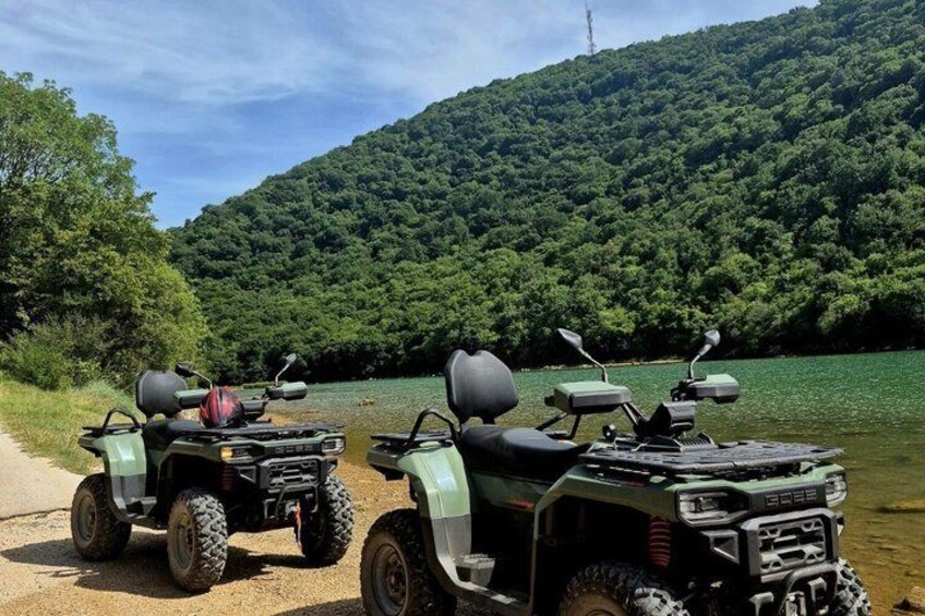 Two off-road vehicles parked by a river with a forested hill in the background. 1