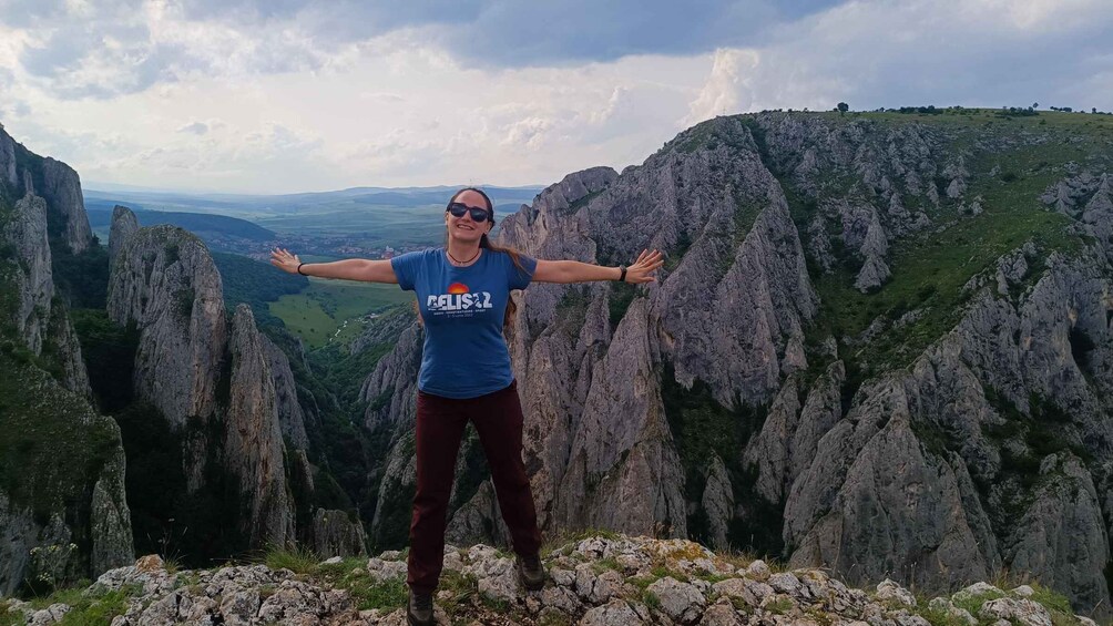 A person standing on a rocky outcrop with arms outstretched, overlooking a valley with steep cliffs and a cross on a hilltop. 5