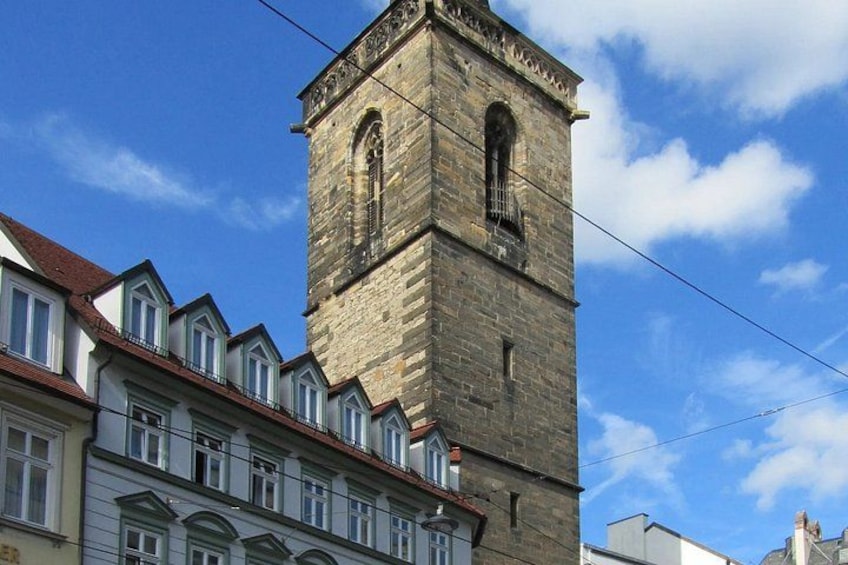A historic stone tower with arched windows and a clock, adjacent to modern buildings with a red roof. 3
