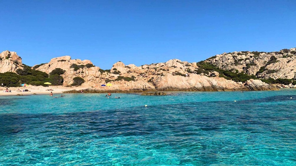 A clear turquoise sea, rocky cliffs, and a beach with people swimming and sunbathing. 4