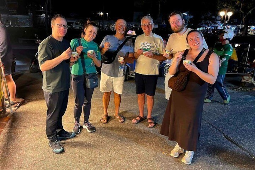 A group of people standing on a sidewalk at night, holding food and drinks. 2