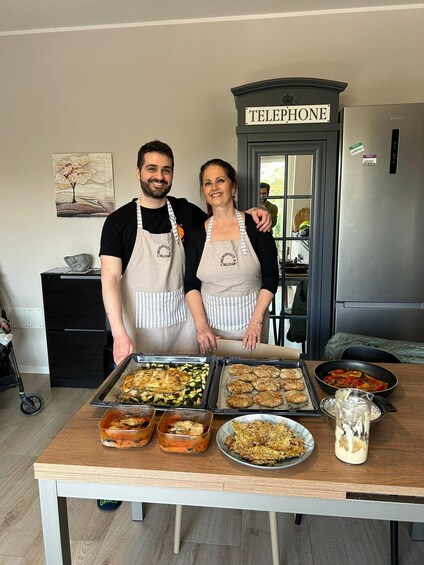 Two people in aprons standing in a kitchen with a table full of dishes. 5