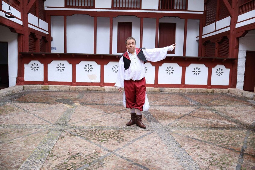 A person in traditional attire standing in a courtyard with a patterned floor and a building with red and white trim in the background. 5