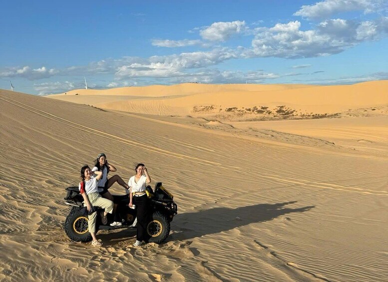 Three people on a dune buggy in a desert landscape with sand dunes and wind turbines in the distance. 4