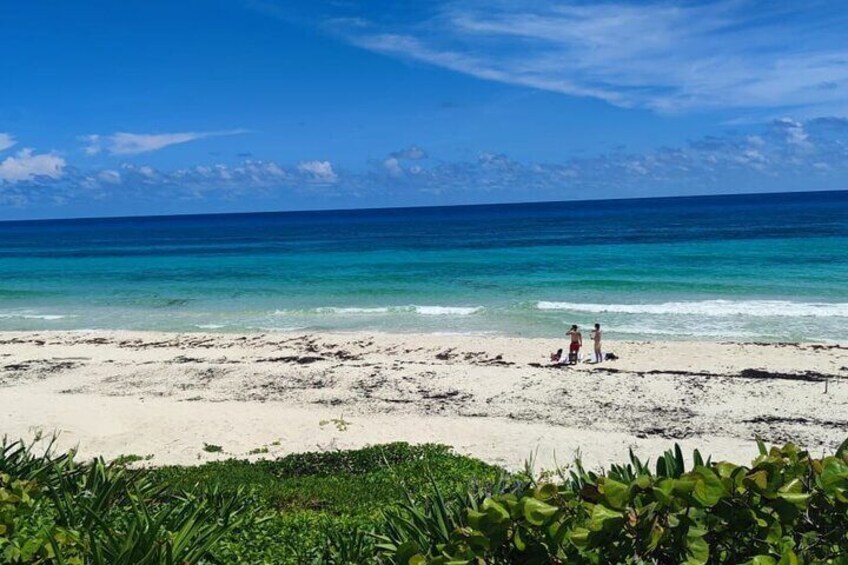 A beach with clear turquoise water, two people standing on the sand, and green vegetation in the foreground. 2