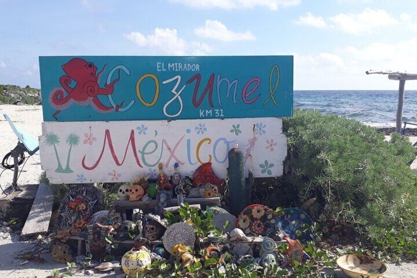 A beach sign with 'El Mirador Ozumel' and 'Mexico' written on it, surrounded by various decorative items and plants. 4