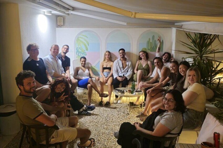 A group of people seated in a room with a tiled floor, some standing, and a cactus plant in the background. 5