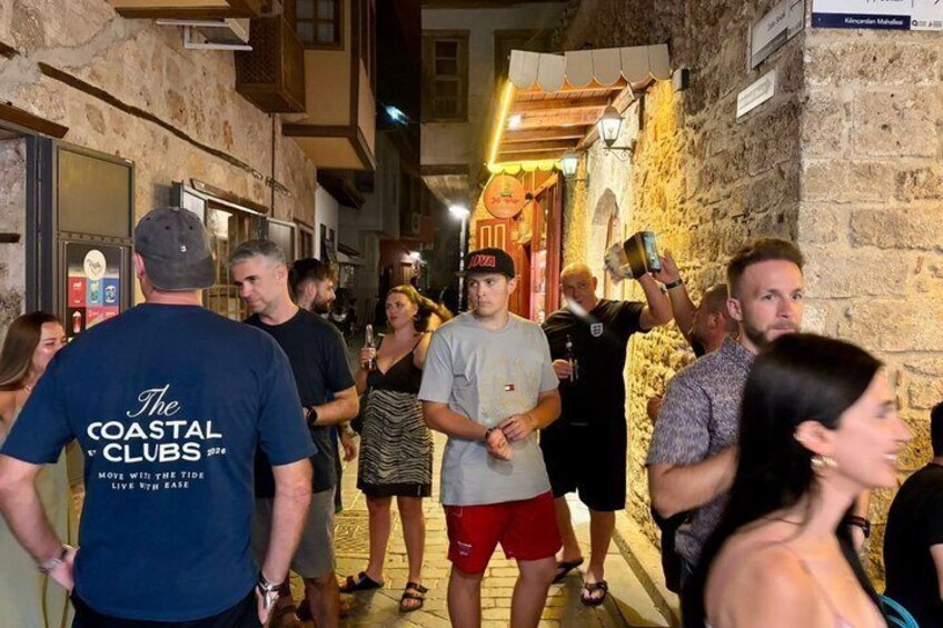 A group of people walking on a narrow street with stone buildings and a sign for 'The Coastal Clubs.' 2