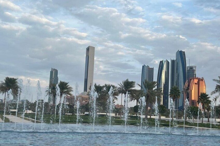 A cityscape with modern skyscrapers, a central tall building, and a fountain area with palm trees. 2