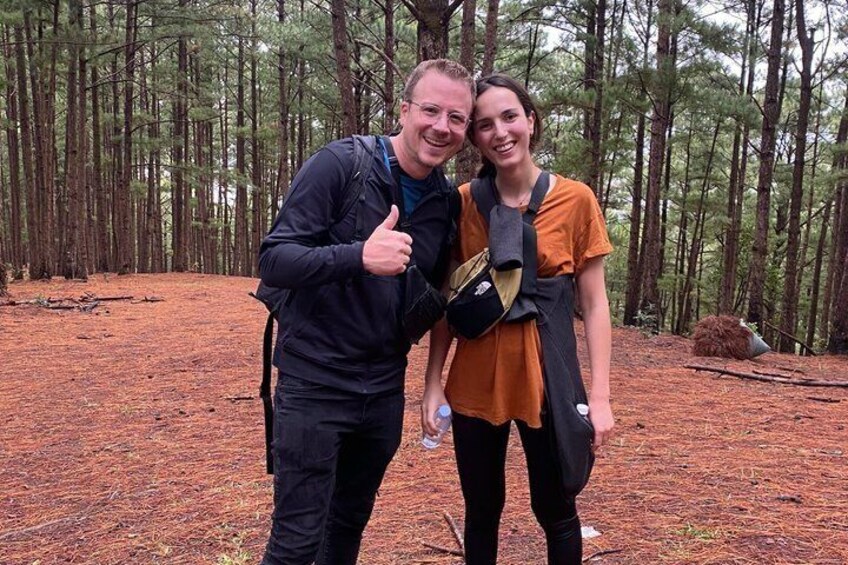 Two people in a forest with a dense tree line and a ground covered in pine needles. 2
