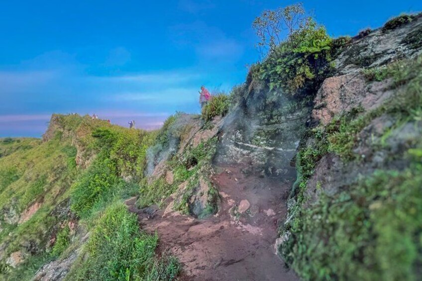 A rocky trail with vegetation, a person standing on a cliff, and a clear sky. 4
