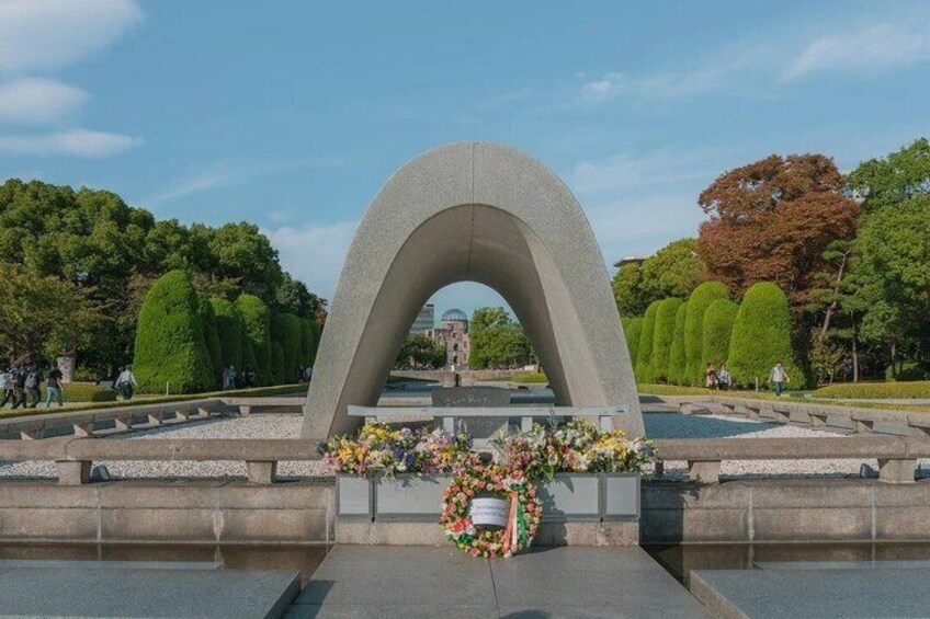 A stone archway with a floral wreath at its base, surrounded by a well-maintained park with trimmed hedges and a clear sky. 2