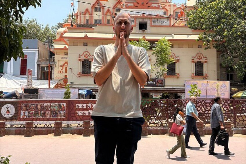 A person in a white shirt with hands clasped in prayer, standing in front of a building with traditional architecture. 2