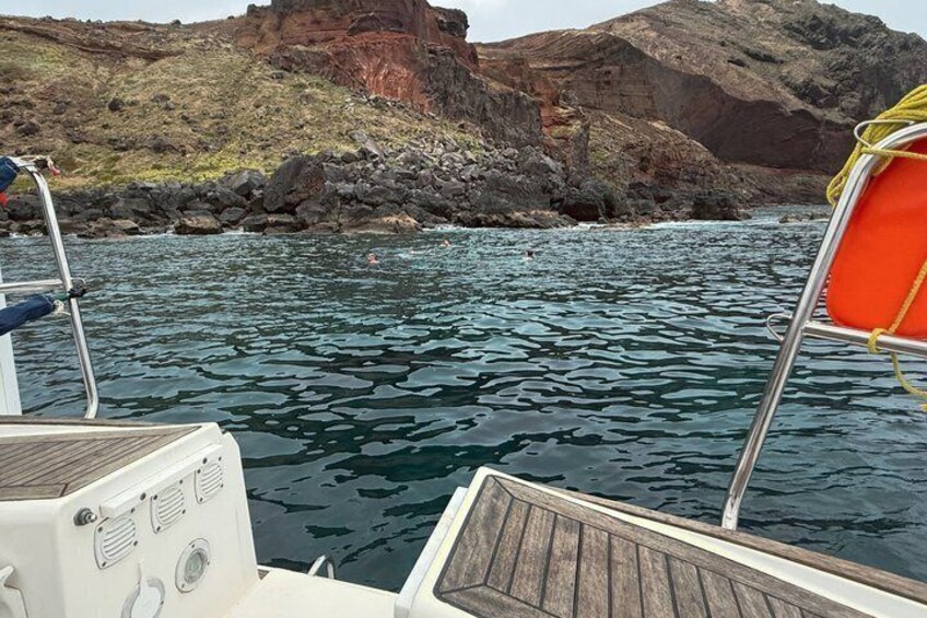 A boat's deck with an orange life preserver, rocky coastline, and calm water. 2