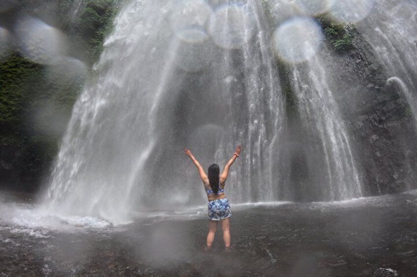 A person standing in water with arms raised, facing a waterfall. 3