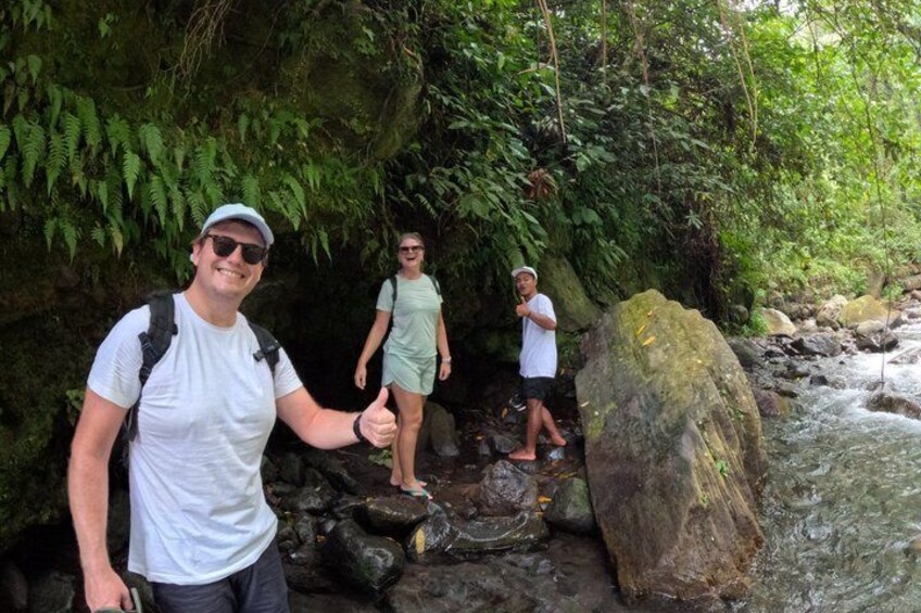 A person giving a thumbs-up gesture, standing on a rocky path with a stream in the background, surrounded by dense green foliage. 5