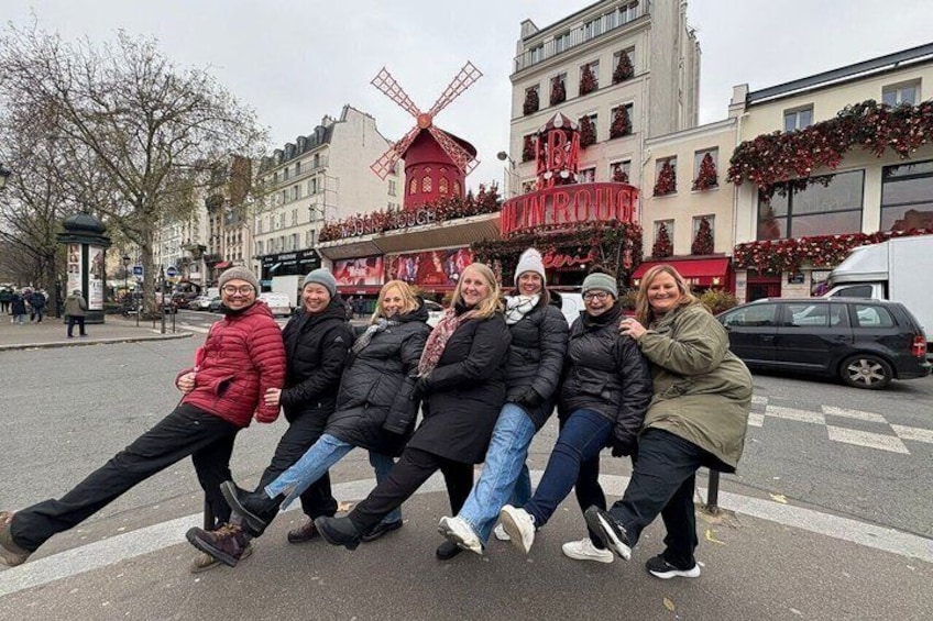 A group of people posing in a city square with a windmill and a building with a sign that reads 'L'ARBRE.' 1