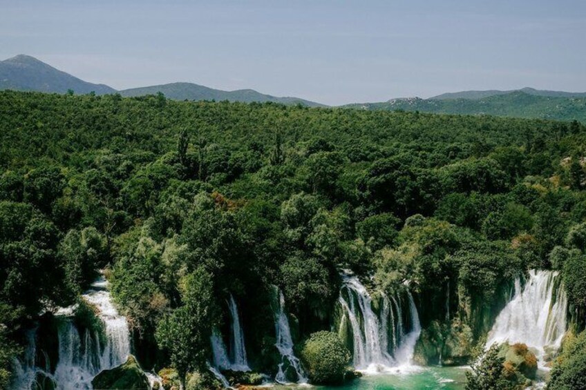 A waterfall cascading through a dense forest with a river below and mountains in the background. 3
